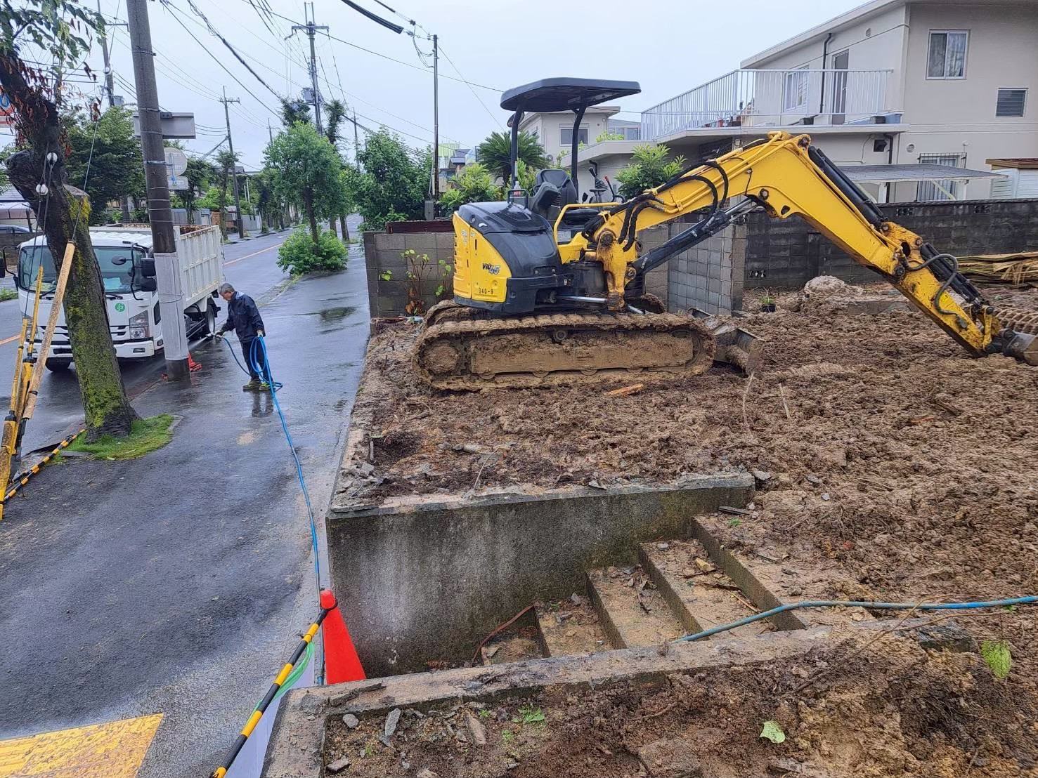 整地中の現場。重機が作業し、作業員が道路側で散水している
